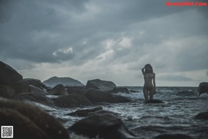 A naked woman standing in the water near some rocks.